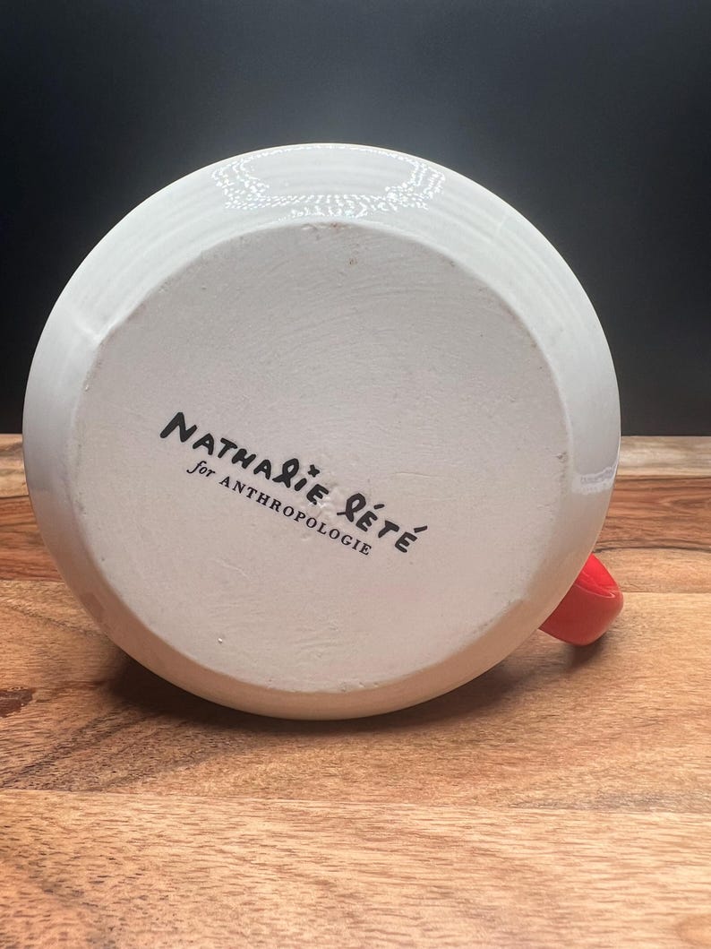 May include: White ceramic mug with a red handle. The bottom of the mug features the text "Nathalie Lete for Anthropologie" in black script. The mug is set on a wooden surface.