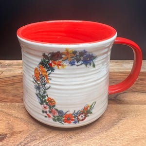 May include: White ceramic mug with a red interior and handle. The mug features a floral design with orange, yellow, and blue flowers, and a small clock. The exterior has a textured, ridged pattern.