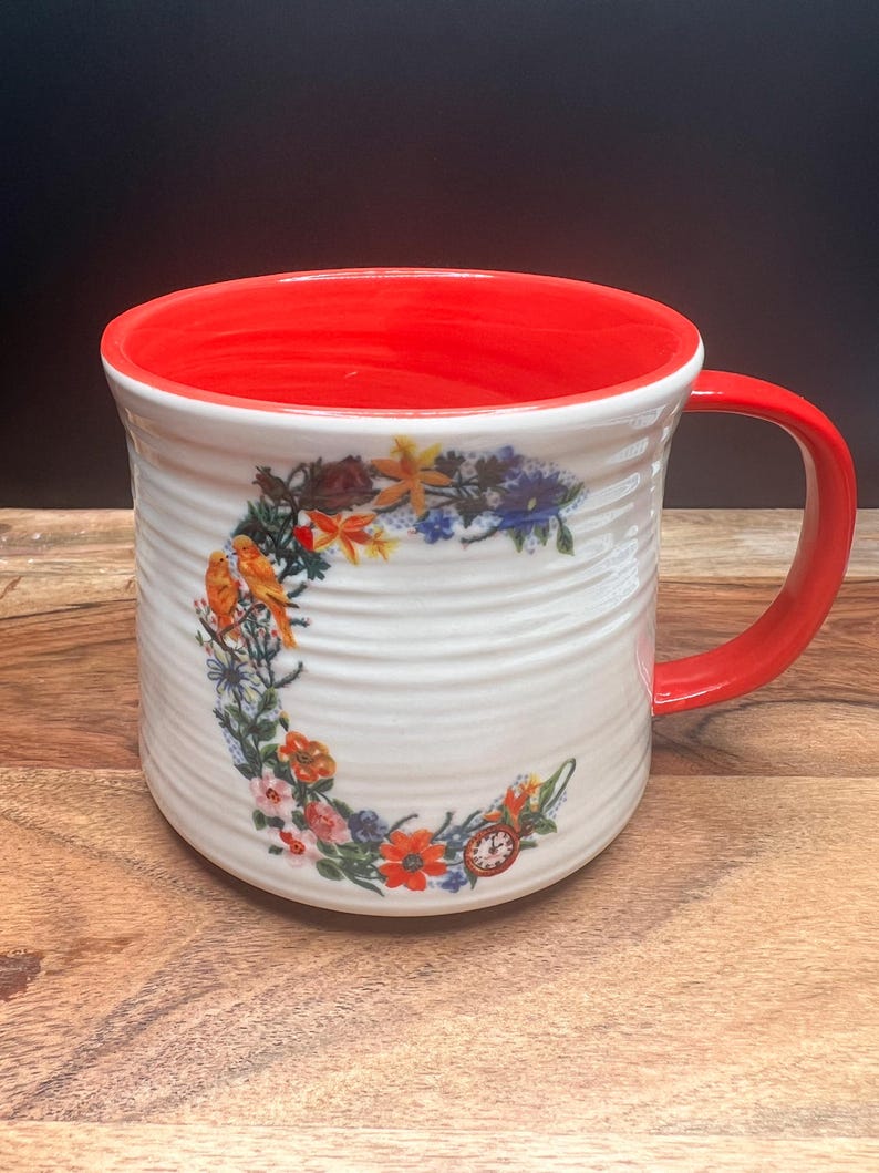 May include: White ceramic mug with a red interior and handle. The mug features a floral design with orange, yellow, and blue flowers, and a small clock. The exterior has a textured, ridged pattern.