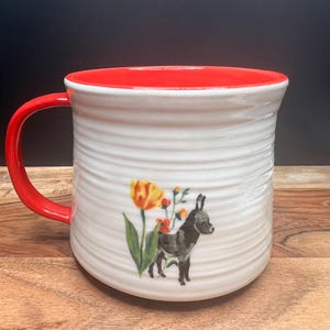 May include: White ceramic mug with a red interior and handle. The mug features a whimsical illustration of a donkey and flowers. The mug has a textured, ridged exterior.