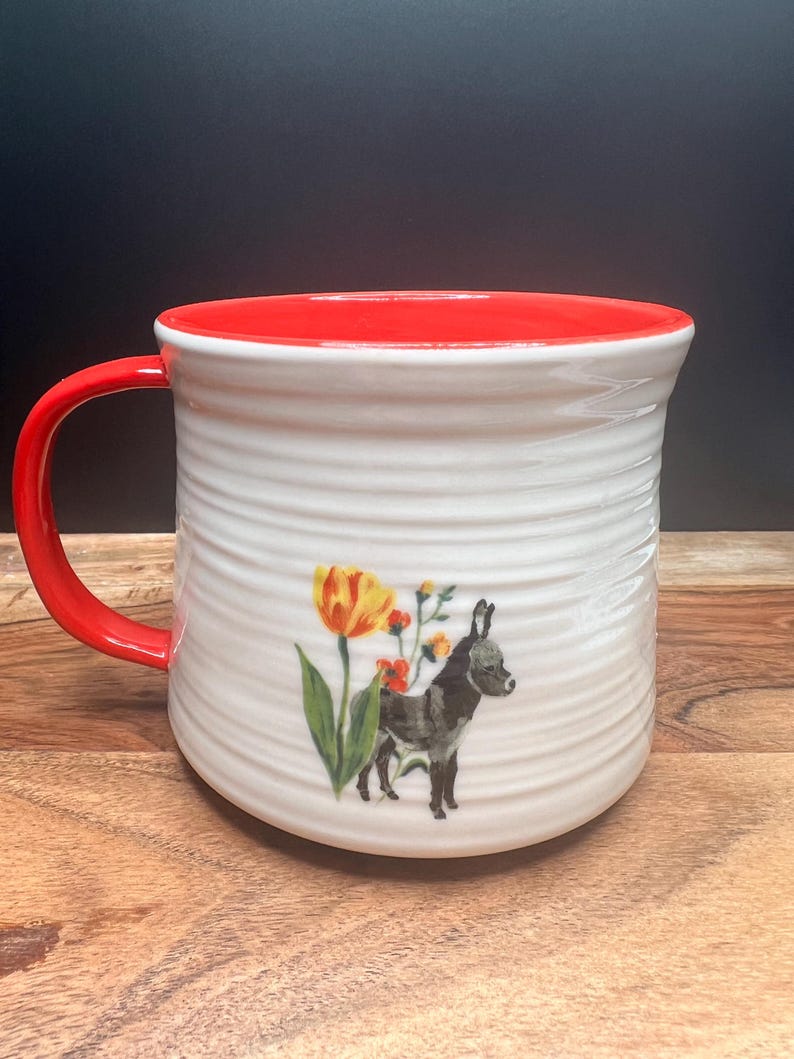 May include: White ceramic mug with a red interior and handle. The mug features a whimsical illustration of a donkey and flowers. The mug has a textured, ridged exterior.