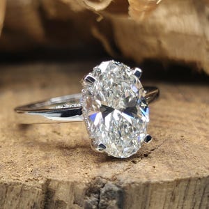 以下が含まれることがあります： オーバルカットのダイヤモンドが特徴の婚約指輪。シルバーのバンドで、木製の表面に置かれています。透明で輝くダイヤモンドが光を反射しています。ジュエリーのクローズアップ。