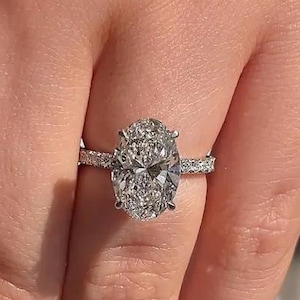 Op de afbeelding: Een ovale diamanten verlovingsring met een pavé band. De ring heeft een grote, sprankelende diamant in een prong setting. De band is gemaakt van wit metaal en is versierd met kleine diamanten.