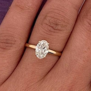 Op de afbeelding: Een gouden ring met een ovaal geslepen diamant. De ring zit aan een vinger met gelakte nagels. Een paarse zeshoekige ringdoos staat op de achtergrond. De band van de ring is dun en geel.