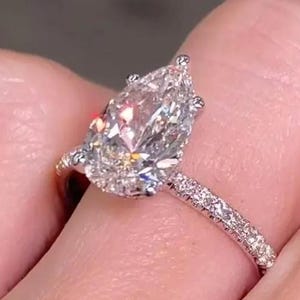 以下が含まれることがあります： 洋梨型のダイヤモンド婚約指輪。大きな透明な洋梨カットのダイヤモンドが、小さなダイヤモンドがちりばめられたバンドにセットされています。指輪は人の手で持たれており、指輪の輝きとデザインが際立っています。