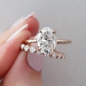 Può includere: Un anello di fidanzamento in oro rosa con un diamante taglio ovale e una fede nuziale abbinata con piccoli diamanti rotondi. Gli anelli sono presentati su uno sfondo morbido e neutro, evidenziando la brillantezza delle pietre.