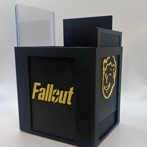 May include: Black box with the word "Fallout" in gold lettering. The box has a clear plastic divider and a gold-colored Vault Boy logo on the side. The box is designed to hold items.