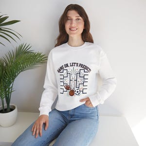 Puede incluir: Sudadera blanca con el texto "GAME ON, LET'S PREDICT!" y un gráfico de un balón de fútbol americano, baloncesto y fútbol. La sudadera tiene mangas largas y cuello redondo.