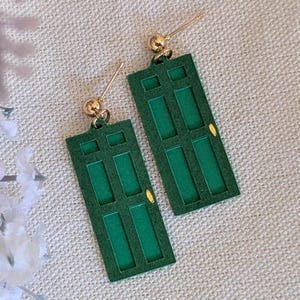 May include: A pair of green door-shaped earrings with gold-colored studs. The earrings have a rectangular shape with three rectangular panels and a small gold accent. The background includes purple and white flowers.