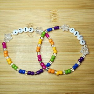 May include: Two beaded bracelets with a rainbow color scheme. One bracelet has the numbers "1312" on white beads. The bracelets are made with clear, colorful, and star-shaped beads. The bracelets are on a wooden surface.