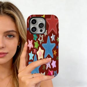May include: A maroon phone case featuring a pattern of colorful stars in various sizes and textures. The stars include red, blue, pink, and metallic silver, with a denim-like blue star prominently displayed. The case is held by a person.