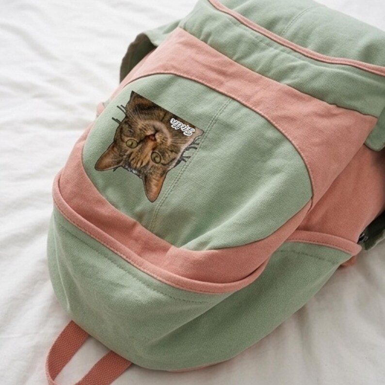 K&ouml;nnte beinhalten: Ein Rucksack aus salbeigr&uuml;nem und altrosa Segeltuch mit einem quadratischen Aufn&auml;her auf der Vordertasche. Der Aufn&auml;her zeigt einen Fotodruck des Gesichts einer braunen getigerten Katze mit dem wei&szlig;en Schreibschrifttext Stella auf der Stirn.