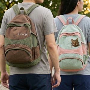 May include: A two-tone olive green and brown canvas backpack with the name George printed in white on the front, and a light green and pink canvas backpack featuring a graphic patch of a brown tabby cat on the front pocket.