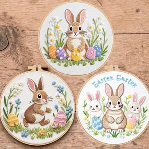 May include: Three embroidered Easter hoop art pieces. Each features a bunny, colourful eggs, and floral accents. One has the text "Easter Easter". The bunnies are brown and white, with the eggs in various colours.