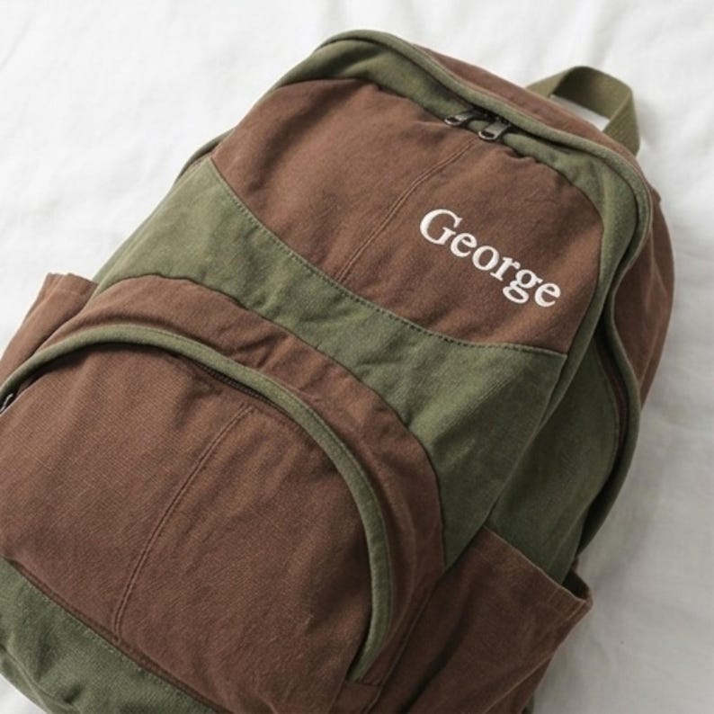 K&ouml;nnte beinhalten: Ein Rucksack aus Segeltuch mit einem farblich abgesetzten Design in Braun und Olivgr&uuml;n. Auf der Vorderseite ist der Name George in wei&szlig;en Buchstaben eingestickt. Die Tasche verf&uuml;gt &uuml;ber einen Rei&szlig;verschluss oben, eine Fronttasche und einen Tragegriff.