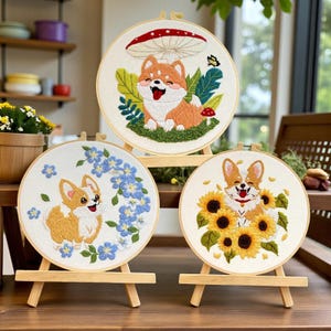 May include: Three embroidered hoops featuring cartoon dogs. One has a mushroom and butterfly, another has blue flowers, and the third has sunflowers. Each hoop is displayed on a small wooden easel, showcasing the detailed needlework.