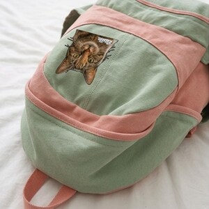K&ouml;nnte beinhalten: Ein Rucksack aus salbeigr&uuml;nem und altrosa Segeltuch mit einem aufgedruckten Bild eines braunen getigerten Katzengesichts. Die Katzengrafik enth&auml;lt den wei&szlig;en kursiven Schriftzug Stella in der N&auml;he des rechten Ohrs. Der Rucksack hat eine Vordertasche.