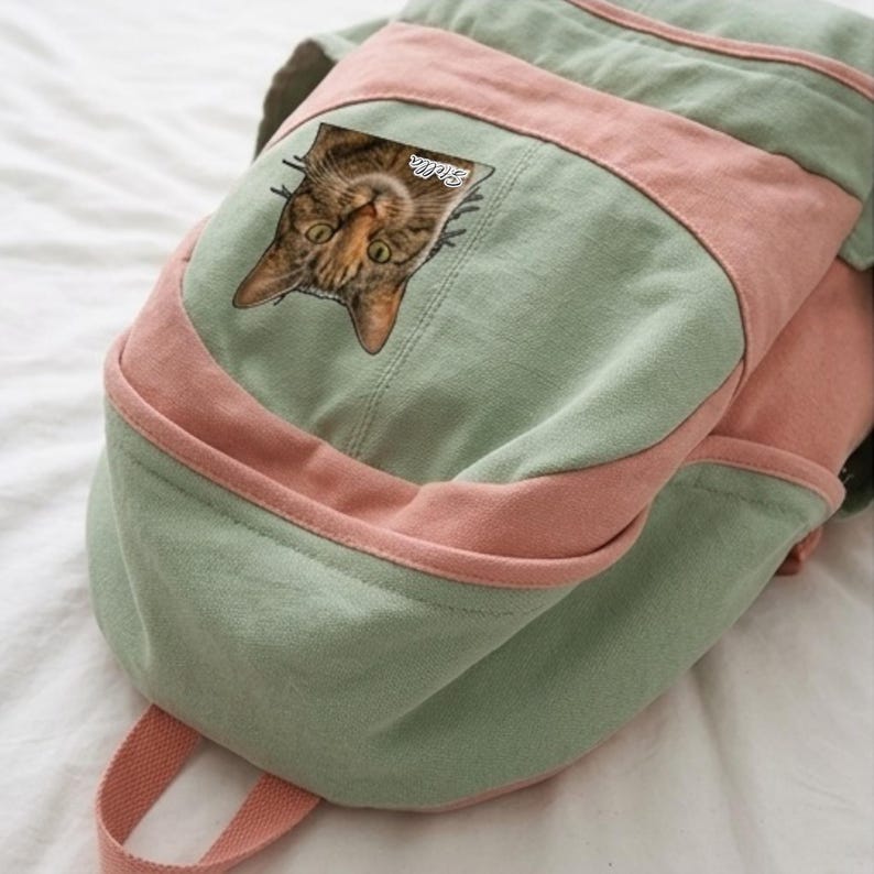 K&ouml;nnte beinhalten: Ein Rucksack aus salbeigr&uuml;nem und altrosa Segeltuch mit einem aufgedruckten Bild eines braunen getigerten Katzengesichts. Die Katzengrafik enth&auml;lt den wei&szlig;en kursiven Schriftzug Stella in der N&auml;he des rechten Ohrs. Der Rucksack hat eine Vordertasche.
