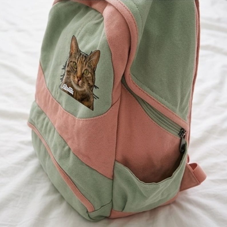 K&ouml;nnte beinhalten: Ein Rucksack aus salbeigr&uuml;nem und altrosa Canvas mit einem aufgen&auml;hten Bild einer braunen getigerten Katze, unter deren Kinn der Name Stella in wei&szlig;er Schreibschrift steht. Der Rucksack verf&uuml;gt &uuml;ber eine Vordertasche, Seitentaschen und einen Rei&szlig;verschluss.