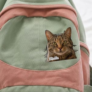 K&ouml;nnte beinhalten: Ein salbeigr&uuml;ner und altrosa Rucksack aus Segeltuch mit einem rechteckigen Aufn&auml;her, der das realistische Portr&auml;t einer braunen getigerten Katze zeigt, wobei der Name Stella in wei&szlig;er Schreibschrift unter dem Kinn steht.