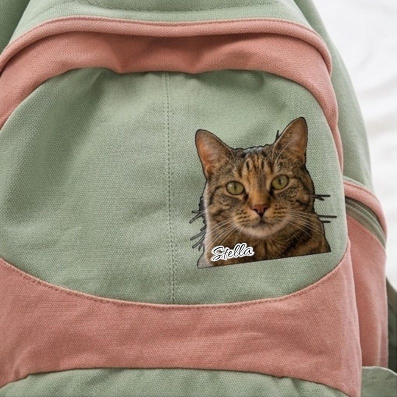 K&ouml;nnte beinhalten: Ein salbeigr&uuml;ner und altrosa Rucksack aus Segeltuch mit einem rechteckigen Aufn&auml;her, der das realistische Portr&auml;t einer braunen getigerten Katze zeigt, wobei der Name Stella in wei&szlig;er Schreibschrift unter dem Kinn steht.