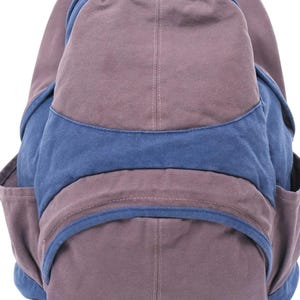 K&ouml;nnte beinhalten: Ein Rucksack aus Segeltuch mit einem Color-Block-Design in gedecktem Violett und Dunkelblau. Die Tasche verf&uuml;gt &uuml;ber ein gro&szlig;es Hauptfach, eine geschwungene Vordertasche mit blauem Besatz und Seitentaschen, alles mit sichtbaren N&auml;hten und Stoffbahnen.