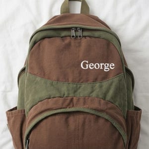 K&ouml;nnte beinhalten: Ein Rucksack aus braunem und olivgr&uuml;nem Segeltuch mit einem Tragegriff, zwei Rei&szlig;verschlussf&auml;chern und Seitentaschen. Das Wort George ist in wei&szlig;em Garn auf das obere Vorderteil der Tasche gestickt, die ein strukturiertes Design aufweist.