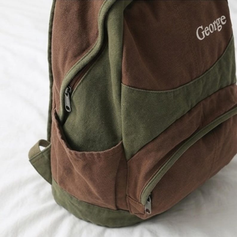 K&ouml;nnte beinhalten: Ein brauner und olivgr&uuml;ner Rucksack aus Segeltuch mit einer Seitentasche mit Rei&szlig;verschluss und dem Namen George, der in wei&szlig;em Garn auf dem oberen Vorderteil eingestickt ist. Die Tasche verf&uuml;gt &uuml;ber ein vorderes Rei&szlig;verschlussfach.