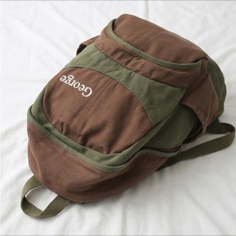 K&ouml;nnte beinhalten: Ein Rucksack aus braunem und olivgr&uuml;nem Segeltuch mit einem abgerundeten oberen Fach und einer Vordertasche. Das Wort George ist mit wei&szlig;em Garn auf den oberen braunen Bereich gestickt. Die Tasche hat olivgr&uuml;ne Bes&auml;tze, Rei&szlig;verschl&uuml;sse und einen Tragegriff.
