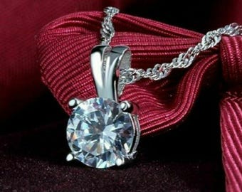 May include: A silver pendant necklace featuring a large, round, clear gemstone. The gemstone is set in a silver metal setting and hangs from a delicate silver chain. The necklace is displayed on a dark red fabric.