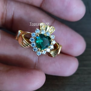 Peut inclure: Une bague Claddagh dorée avec une pierre précieuse verte émeraude en forme de cœur, entourée de petites pierres bleues claires et blanches. La conception de la bague comprend deux mains tenant une couronne au-dessus du cœur. La bague est tenue dans une main.