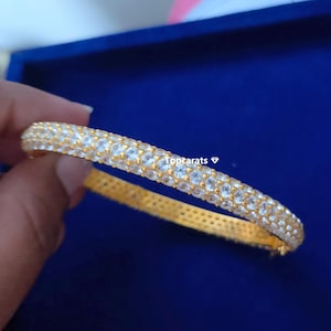 May include: A gold-coloured bangle bracelet set with many small, clear gemstones. The bracelet is held against a dark blue background, highlighting its sparkle. The text "topcarats" is visible.