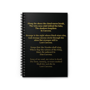 May include: Black spiral-bound notebook with gold text. The text includes the phrases "Along the shore the cloud waves break" and "Lost Carcosa." The notebook has a black cover and white pages. The notebook is designed for writing.