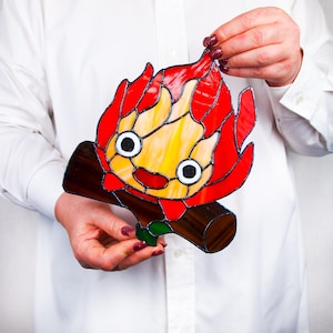 May include: Stained glass artwork depicting a cartoon fire character with red and yellow flames, black eyes, and a smiling mouth. The character is perched on a brown log with green accents. The artwork is held up against a white background.