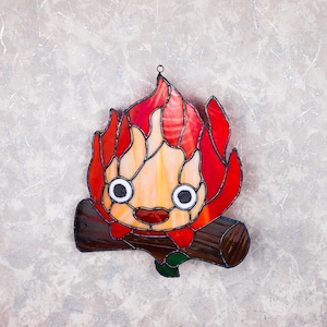 May include: Stained glass artwork depicting a cartoonish fire character with a smiling face and large eyes. The flames are red and orange, with a brown log base. The piece is designed to hang, with a small metal loop at the top.