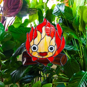 May include: Stained glass artwork depicting a cartoon fire character with red and yellow flames, black eyes, and a smiling red mouth. The character is perched on a brown log, with a metal loop for hanging. The background is lush green foliage.