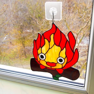 May include: A stained glass suncatcher featuring a cartoon-style fire character with red and yellow flames, black eyes, and a smiling mouth. The character is perched on a brown log with a green leaf.