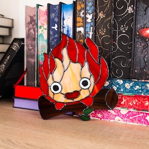 May include: A stained glass art piece depicting a cartoon flame character with a smiling face, red flames, and a log base. The artwork is set against a backdrop of colorful books, creating a whimsical and decorative scene.
