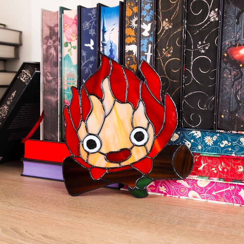 May include: A stained glass art piece depicting a cartoon flame character with a smiling face, red flames, and a log base. The artwork is set against a backdrop of colorful books, creating a whimsical and decorative scene.