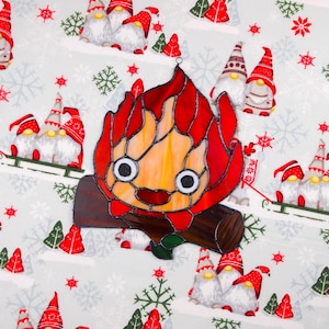 May include: A stained glass decoration in the shape of a cartoon campfire with a smiling face. The flames are red and orange, and the log is brown. The background is a light gray fabric with gnomes, snowflakes, and Christmas trees.