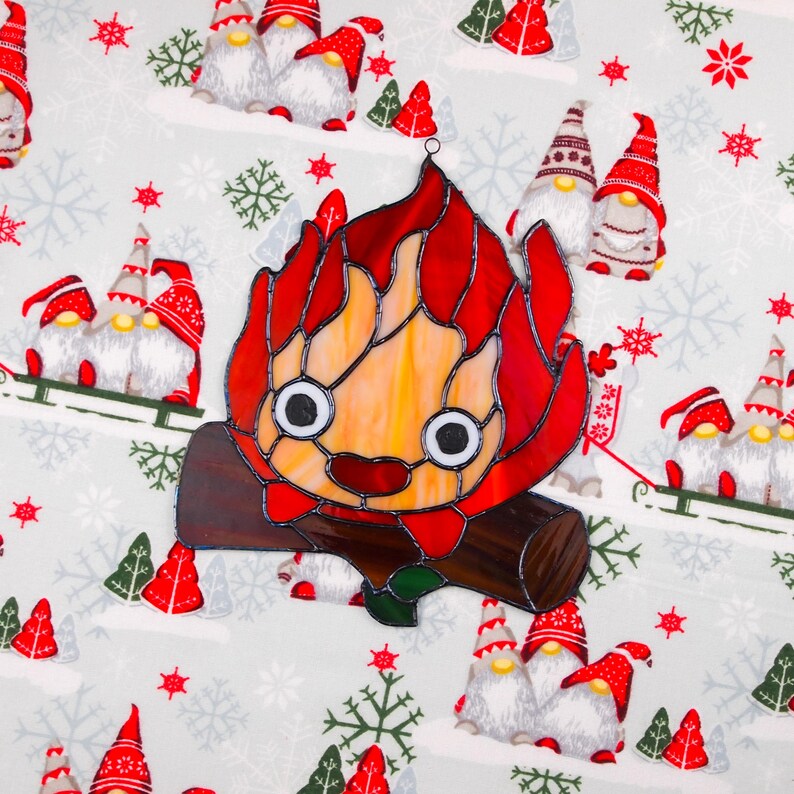May include: A stained glass decoration in the shape of a cartoon campfire with a smiling face. The flames are red and orange, and the log is brown. The background is a light gray fabric with gnomes, snowflakes, and Christmas trees.