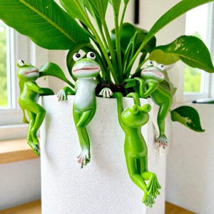 Puede incluir: Una maceta de cerámica blanca con cuatro figuras de ranas verdes colgando de los lados. Las ranas tienen vientres blancos y están posicionadas como si estuvieran trepando o colgando de la maceta. La maceta contiene una planta verde.