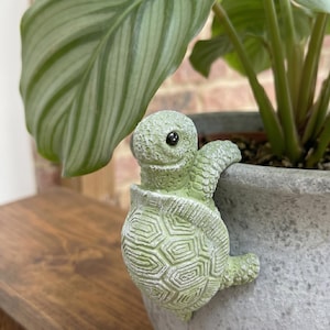 Puede incluir: Una figura de tortuga verde claro posada en el borde de una maceta gris. La tortuga tiene un caparazón texturizado y una cara sonriente con ojos negros. Una gran hoja verde de planta es visible en el fondo.