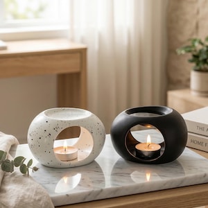 Op de afbeelding: Twee bolvormige kaarshouders. Eén is wit met zwarte spikkels, de andere is zwart. Beide hebben een grote cirkelvormige opening en een brandende waxinelichtkaars erin. De kaarshouders staan op een marmeren oppervlak.