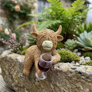May include: A brown Highland cow ornament holding a glass of red wine, set in a stone planter with plants. The cow has horns and a light green nose. The wine glass is clear, and the wine is a deep red colour.