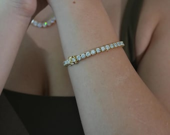 Op de afbeelding: Een gouden armband met ronde, heldere edelstenen. De armband is vastgemaakt met een sluiting. Een bijpassende ketting is zichtbaar op de achtergrond. De sieraden worden om een arm gedragen.