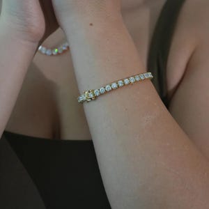 May include: A gold bracelet set with clear, round gemstones. The bracelet is secured with a clasp. A matching necklace is visible in the background. The jewellery is worn on an arm.