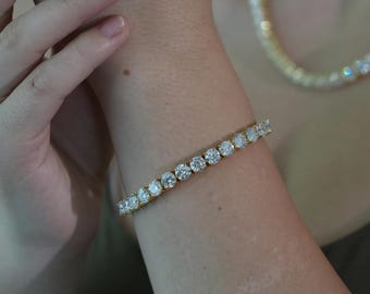Op de afbeelding: Een gouden armband met een rij ronde, heldere edelstenen. De armband wordt om een pols gedragen, en een bijpassende ketting is zichtbaar op de achtergrond. De sieraden zijn geplaatst tegen een neutrale achtergrond.