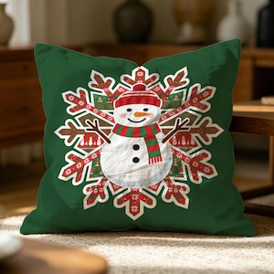 Può includere: Un cuscino quadrato di colore verde bosco intenso con una grafica centrale raffigurante un pupazzo di neve bianco che indossa un cappello rosso e una sciarpa a righe rosse e verdi, posizionato sopra un grande e intricato motivo a fiocco di neve.
