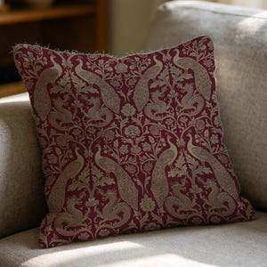 May include: A square throw pillow featuring a deep burgundy background with a repeating pattern of beige peacocks and mythical creatures intertwined with floral and leafy motifs. The fabric has a textured appearance with visible woven details.